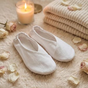 May include: A pair of white linen slippers with a soft, textured appearance. The slippers are placed on a fluffy, cream-colored surface, alongside a lit candle, a folded blanket, and rose petals.