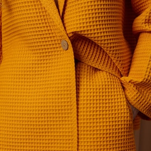 May include: Mustard yellow waffle-weave blazer and shorts set. The blazer features a single wooden button closure and a notched lapel. The fabric has a textured, grid-like pattern. The shorts are visible at the bottom of the image.