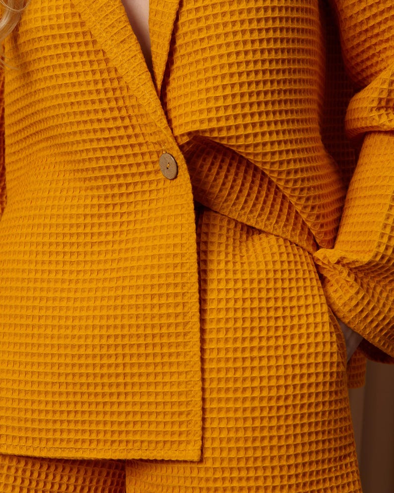 May include: Mustard yellow waffle-weave blazer and shorts set. The blazer features a single wooden button closure and a notched lapel. The fabric has a textured, grid-like pattern. The shorts are visible at the bottom of the image.