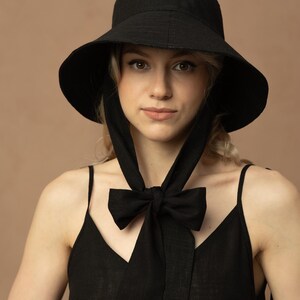 May include: A black sun hat with a wide brim and a matching bow tie. The hat has long ties that hang down and are tied in a bow. The woman is wearing a black tank top.