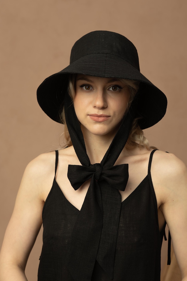 May include: A black sun hat with a wide brim and a matching bow tie. The hat has long ties that hang down and are tied in a bow. The woman is wearing a black tank top.