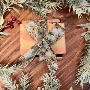 May include: A bar of handmade soap with a peach and cream color gradient, resting on a wooden soap dish. The soap is adorned with a decorative ribbon featuring pine cones and evergreen branches. The image is framed with festive greenery.