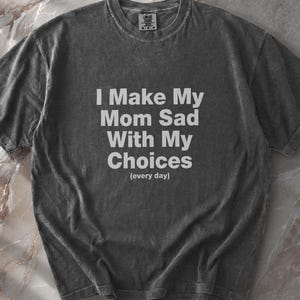May include: A charcoal grey short-sleeved t-shirt with a washed-out texture. The front features white text that reads: I Make My Mom Sad With My Choices (every day). The shirt has a crew neckline and a standard fit with visible fabric folds.