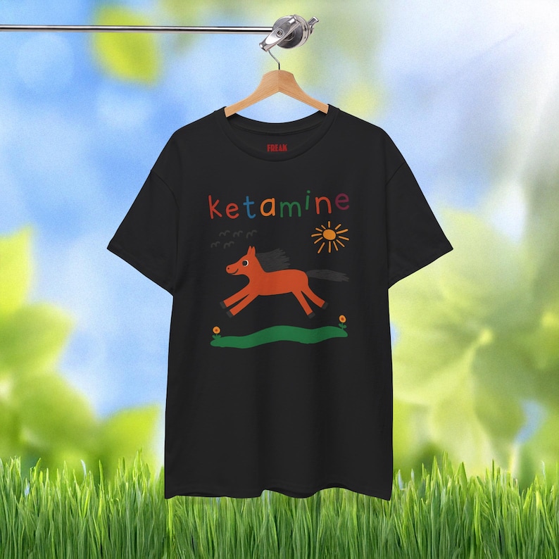 Op de afbeelding: Zwart T-shirt met een kleurrijke afbeelding van een paard dat in een veld rent met een zon en het woord "ketamine" in regenboogkleuren.