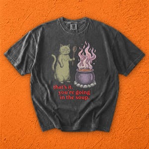 Puede incluir: Una camiseta gris carbón con un gráfico de un gato sosteniendo una cuchara de madera junto a un caldero morado burbujeante con vapor rosa. Debajo de la ilustración, el texto rojo dice: that's it. you're going in the soup.