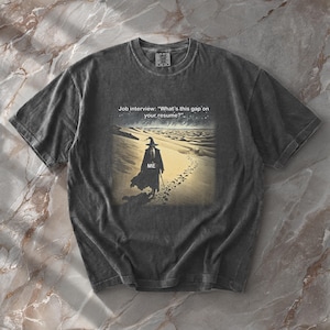May include: A charcoal gray short-sleeve t-shirt featuring a graphic of a wizard walking across a desert. The text above reads, Job interview: "What's this gap on your resume?" and the word ME is printed on the wizard's cloak, showing a trail of footprints.