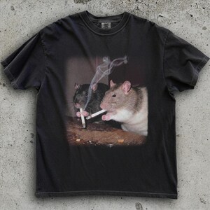 Puede incluir: Camiseta gris oscuro con un gr&aacute;fico de dos ratas fumando cigarrillos. Una rata es negra, la otra es marr&oacute;n y blanca. El humo se eleva de los cigarrillos. La camiseta tiene cuello redondo y mangas cortas.