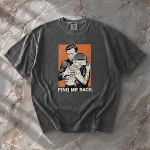 Op de afbeelding: Donkergrijs t-shirt met een retro-afbeelding van twee figuren die elkaar omhelzen, waarvan één met een vintage radio over hun gezicht. De afbeelding staat tegen een oranje achtergrond, met de tekst "PING ME BACK" eronder.
