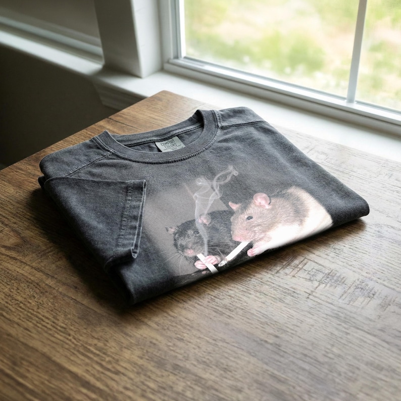 Puede incluir: Camiseta gris oscuro con un gr&aacute;fico de dos ratas, una fumando un cigarrillo. La rata de la derecha es marr&oacute;n claro y blanca, la otra es negra. La camiseta est&aacute; doblada sobre una superficie de madera.