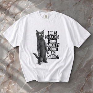 May include: White t-shirt featuring a black cat illustration with the text "Does Shaking From Anxiety Count As Cardio?". The cat is standing upright. The shirt has a crew neck and short sleeves.