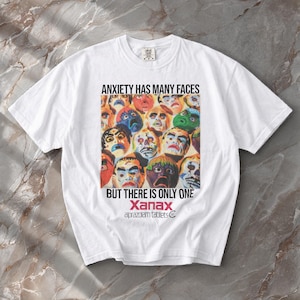 May include: A white short-sleeve t-shirt featuring a graphic print of multiple colorful, expressive faces. Above the graphic is the text ANXIETY HAS MANY FACES and below it is BUT THERE IS ONLY ONE, followed by the text Xanax alprazolam tablets.