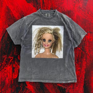 May include: A dark gray t-shirt featuring a square image of a doll with messy blonde hair, smudged makeup, and a neutral expression. The shirt has a crew neck and short sleeves. The background is a textured red.