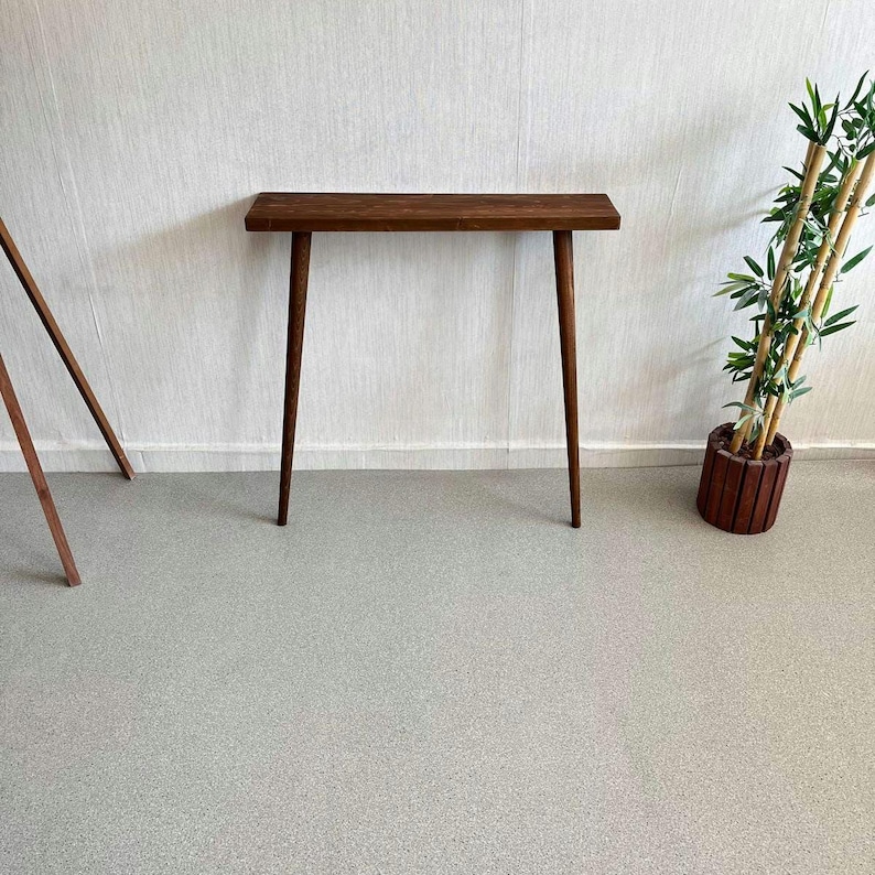 Op de afbeelding: Een console in mid-century modern stijl met een donkerbruine afwerking. De tafel heeft een rechthoekig blad en vier taps toelopende poten. De tafel is ongeveer 122 cm lang. Rechts staat een decoratieve bamboeplant in een houten pot.