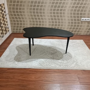 以下が含まれることがあります： 4本の細い黒い脚が付いた、黒い腎臓型のコーヒーテーブル。 テーブルは、花柄の明るい色のラグの上に置かれています。 ラグは木製の床に置かれています。