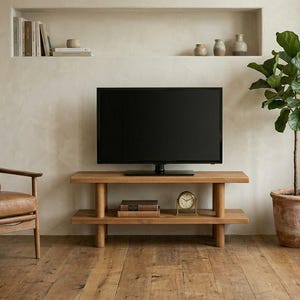 Puede incluir: Salón con un mueble de TV de madera con un televisor de pantalla negra. Un sillón de cuero está a la izquierda y una planta en maceta a la derecha. La habitación tiene una paleta de colores neutros y suelo de madera.