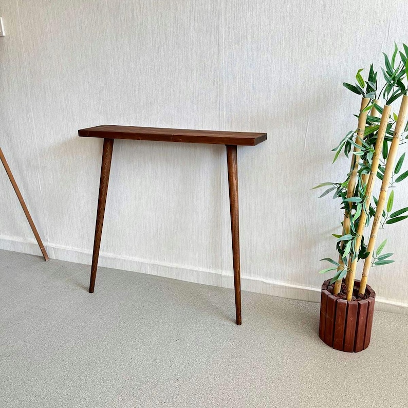 Op de afbeelding: Een console met een afwerking in donkerbruin, in mid-century stijl. De tafel heeft een rechthoekig blad en vier slanke, taps toelopende poten. De tafel is ongeveer 122 cm lang.