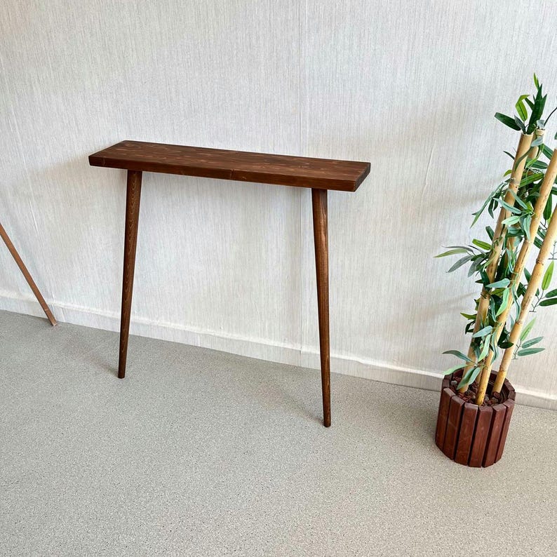 Op de afbeelding: Een donkerbruine houten console met een rechthoekig blad en drie slanke, taps toelopende poten. De tafel is ongeveer 76 cm lang en staat tegen een witte muur. Rechts staat een decoratieve plant in een houten pot.