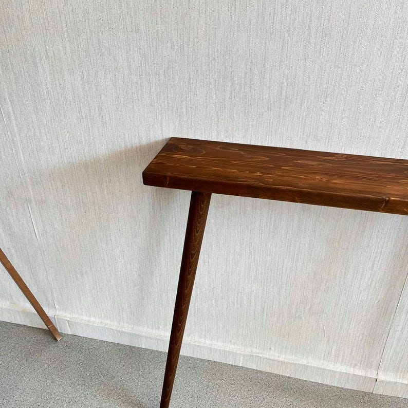 Op de afbeelding: Een lange, smalle houten console met een rijke bruine afwerking. De tafel heeft een rechthoekig blad en drie slanke, schuine poten. De houtnerf is zichtbaar en voegt textuur toe. De tafel is ontworpen om tegen een muur te plaatsen.