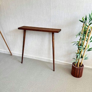 Op de afbeelding: Een smalle, donkerbruine houten consoletafel met een rechthoekig blad en vier slanke, taps toelopende poten. De tafel is ongeveer 122 cm lang en staat tegen een witte muur. Rechts staat een plant in een pot.