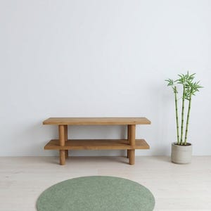 以下が含まれることがあります： 2段の棚と円筒形の脚が付いた、明るい茶色の木製ベンチ。ベンチの前には、丸いセージグリーンのラグが敷かれています。ベンチの右側には鉢植えの竹があります。