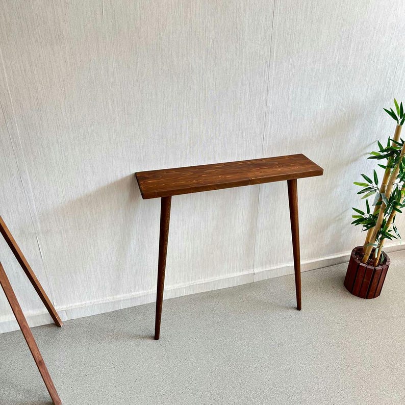Op de afbeelding: Een smalle, rechthoekige houten console met een donkerbruine afwerking. De tafel heeft een eenvoudig ontwerp met vier slanke, taps toelopende poten. De tafel staat tegen een witte muur, met een plant in een pot aan de rechterkant. De tafel is ongeveer 100 cm lang.