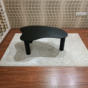 May include: A black, free-form shaped coffee table with rounded edges and three legs. The table is sitting on a white rug with a floral pattern.