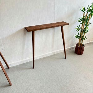 Op de afbeelding: Een smalle houten console met een rechthoekig blad en vier taps toelopende poten. De tafel is gebeitst in een warme bruine kleur en staat tegen een witte muur. Rechts staat een bamboeplant in pot.