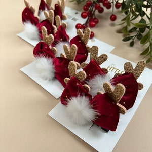 Peut inclure: Ensemble de six pinces à cheveux en velours rouge en forme de renne avec des bois en paillettes dorées et des pompons blancs. Les pinces sont sur une carte blanche avec le texte "Little Bae" dessus.