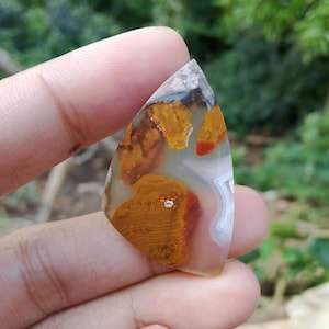 May include: A triangular shaped agate cabochon with a white, brown, and red pattern. The agate has a translucent appearance and is a natural stone.