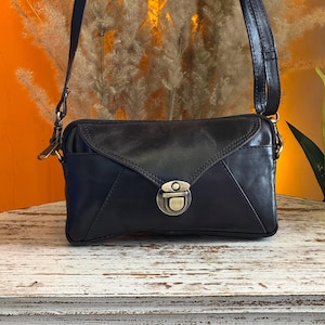 Puede incluir: Bolso bandolera de cuero negro con cierre de solapa y cierre metálico. El bolso tiene una correa larga y ajustable y una forma rectangular. El bolso está sobre una superficie de madera blanca desgastada.