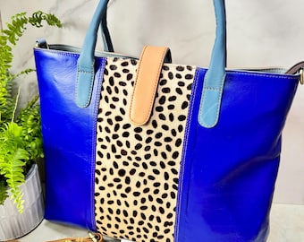 May include: A tote bag featuring a vibrant royal blue exterior with a central vertical panel of beige and black animal print fur. The bag includes light blue leather handles, a tan leather closure strap, and a detachable tan shoulder strap.