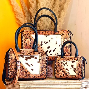 Könnte beinhalten: Drei Lederhandtaschen mit Kuhfell-Einsatz und floralen Verzierungen. Die Taschen haben schwarze Griffe und Riemen. Die größte Tasche befindet sich im Hintergrund, mit einer mittleren und einer kleinen Tasche davor.