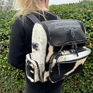Puede incluir: Una mochila de piel de vaca negra y blanca con una solapa de cuero negro y un diseño bordado blanco. La mochila tiene dos bolsillos grandes con cremallera y un bolsillo más pequeño en la parte delantera.