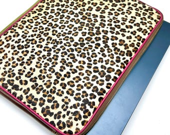 May include: A rectangular laptop sleeve featuring a beige and black leopard print pattern on the front surface. The sleeve is enclosed by a metallic zipper with a bright pink trim along the edges and a silver zipper pull tab at the corner.
