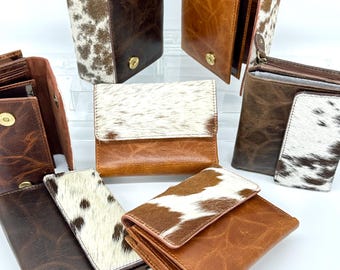 May include: A collection of six leather wallets in various shades of brown, featuring cowhide panels with white and brown spotted patterns. Each wallet includes gold-toned metal snap closures and textured leather surfaces with visible grain details.