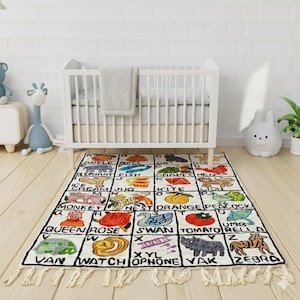 May include: A rectangular alphabet rug with a white background and black borders. Each square features a letter and a corresponding image, such as an apple, elephant, fish, and zebra. The rug has a fringed edge and is placed in a nursery setting.