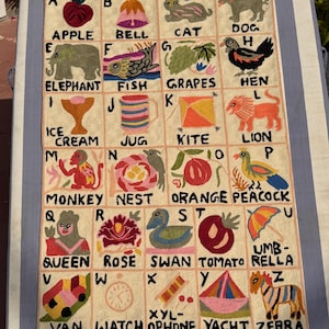 May include: Embroidered alphabet wall hanging with colourful illustrations of objects like an apple, cat, dog, and zebra. The beige fabric has a fringe at the bottom. Letters A to Z are displayed in individual squares.