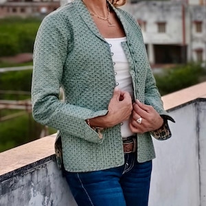 Peut inclure: Veste matelassée vert clair avec un motif texturé. La veste a un col cranté et est boutonnée. La personne porte un haut blanc, un jean bleu et une ceinture marron. Un collier et une bague dorés sont également visibles.