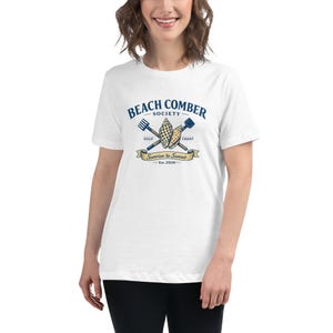 May include: White t-shirt with a "Beach Comber Society" logo in blue and yellow. The logo includes crossed beach tools, seashells, and the text "Sunrise to Sunset - Est. 2026".