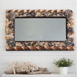 May include: Rectangular mirror with a frame adorned with various seashells and decorative shell flowers. The mirror's frame is dark brown, and the seashells are in shades of brown, beige, and white. The mirror is a decorative coastal home decor item.