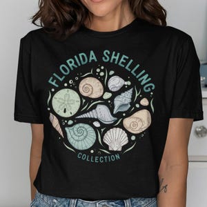 Może przedstawiać: Czarny t-shirt z krótkim rękawem z okrągłą grafiką przedstawiającą różne muszle i sercówkę. Napis "FLORIDA SHELLING" jest wygięty w łuk nad muszlami w kolorze morskim, a słowo "COLLECTION" jest nadrukowane w kolorze morskim poniżej muszli.
