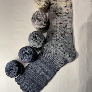 Könnte beinhalten: Eine handgestrickte Socke in Blau-, Grau- und Cremetönen mit einem strukturierten Muster. Fünf Knäuel Garn in passenden Farben liegen neben der Socke. Das Garn und die Socke befinden sich auf einer weißen Oberfläche.