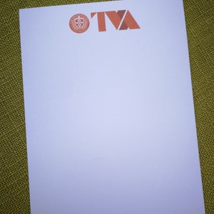 Könnte beinhalten: Weißer Notizblock mit einem roten und weißen Logo, auf dem "TVA" steht, und einem kleineren Logo mit einem Schild und einem Kreuz. Der Text "VARIATION CASE FILE" befindet sich am unteren Rand des Notizblocks.