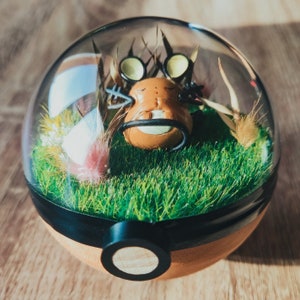 Puede incluir: Un terrario de cúpula de vidrio con una criatura marrón y negra en su interior. La criatura tiene ojos grandes y una boca pequeña. El terrario está lleno de hierba verde y pequeñas flores blancas.