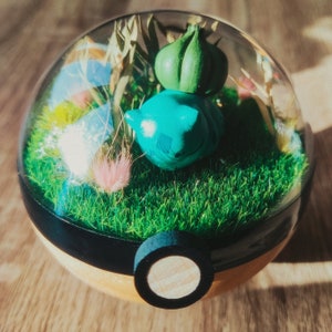 May include: A clear glass sphere with a green and blue figurine inside. The sphere is filled with green grass and a variety of dried flowers. The sphere is sitting on a wooden surface.