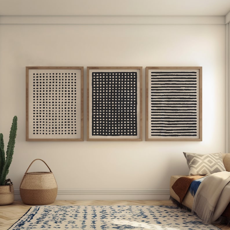 May include: Three framed abstract art prints. The prints feature black and white geometric patterns: a grid, a series of squares, and horizontal lines. The frames are a light brown color, and the art is displayed on a white wall.