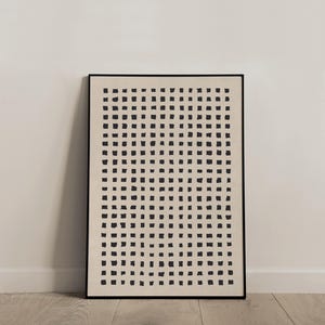 May include: A framed abstract art print featuring a grid of black square shapes on a cream-colored background. The artwork is encased in a black frame and leans against a white wall and a wooden floor.