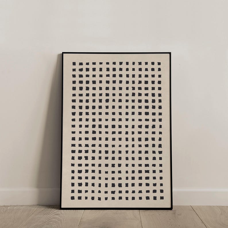 May include: A framed abstract art print featuring a grid of black square shapes on a cream-colored background. The artwork is encased in a black frame and leans against a white wall and a wooden floor.