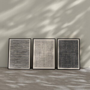 May include: Three framed abstract art prints. The prints feature black and white designs: horizontal lines, a grid of dots, and a grid of squares. The frames are black, and the prints are displayed against a neutral wall and floor.