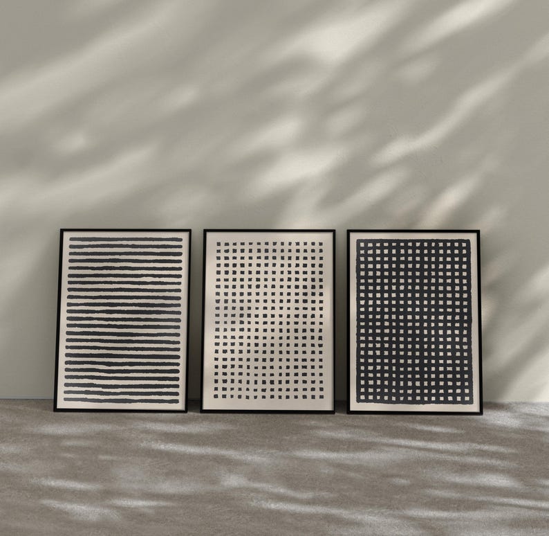 May include: Three framed abstract art prints. The prints feature black and white designs: horizontal lines, a grid of dots, and a grid of squares. The frames are black, and the prints are displayed against a neutral wall and floor.